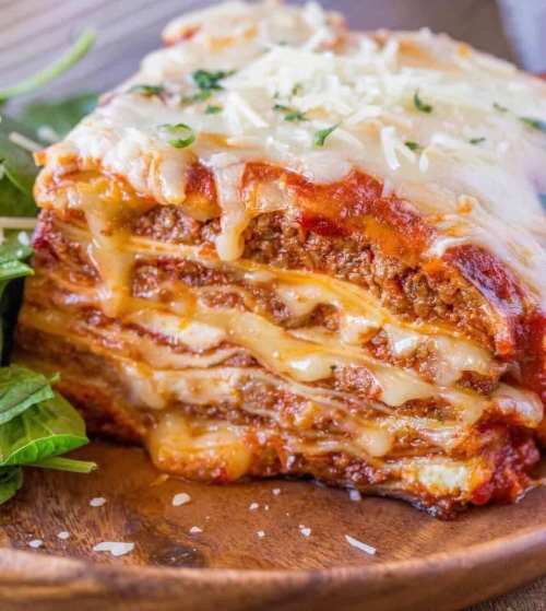A slice of cheesy lasagna with layers of meat sauce and melted cheese, served on a wooden plate with greens.
