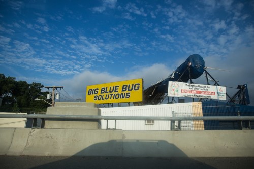 A large billboard reads "BIG BLUE BUG SOLUTIONS" against a cloudy sky, with a blue bug figure on top.
