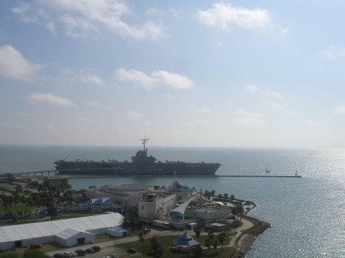 A large aircraft carrier docked near a waterfront, with a clear sky and calm water in the background.