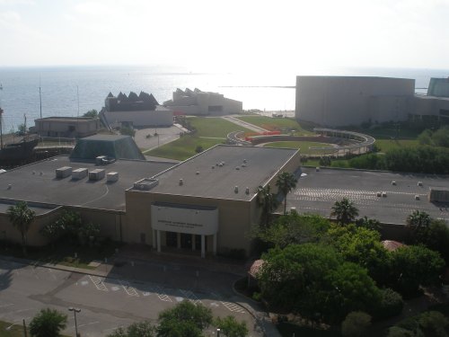 Aerial view of a coastal area featuring buildings, green spaces, and a body of water in the background.