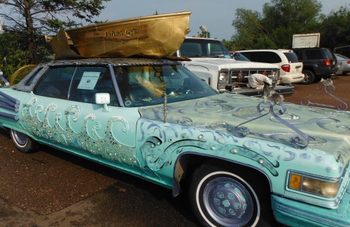 A creatively decorated car with ocean wave designs and a gold boat on top, parked in a lot surrounded by other vehicles.