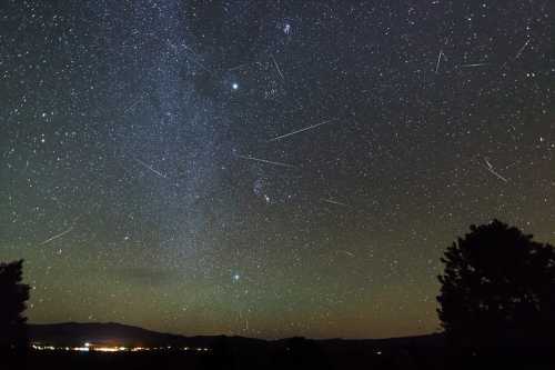 A starry night sky filled with stars and meteor trails, with a distant landscape illuminated by city lights.