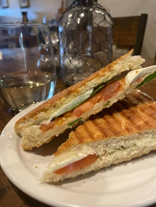 A toasted sandwich with layers of tomato, cheese, and greens, served on a plate next to a glass of white wine.