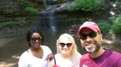 Three people smiling in front of a waterfall and pool, surrounded by lush greenery on a sunny day.