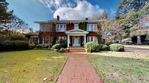 A large red brick house with a front porch, surrounded by greenery and a well-maintained lawn under a blue sky.