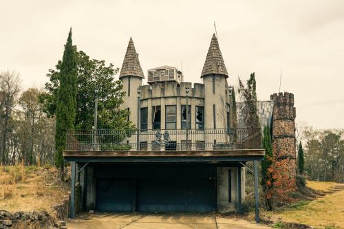 An abandoned castle-like structure with towers, surrounded by overgrown grass and trees, under a cloudy sky.