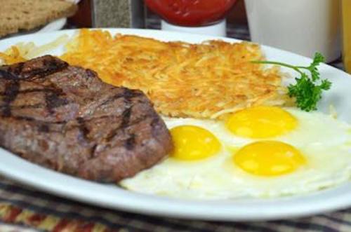 A plate of breakfast featuring grilled steak, three sunny-side-up eggs, and crispy hash browns, garnished with parsley.