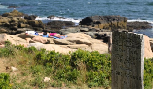A weathered sign marks a coastal path, with people sunbathing on rocks by the ocean in the background.
