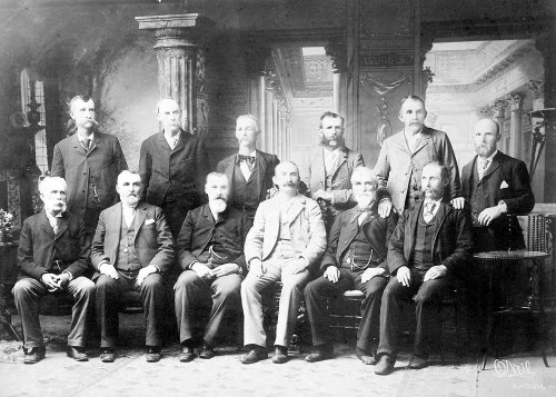 A historic black-and-white photo of twelve men in formal attire, posed in a vintage interior setting.