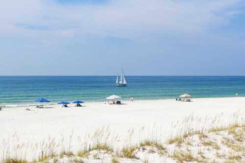 A serene beach scene with white sand, blue umbrellas, and a sailboat on calm waters under a clear sky.
