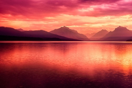 A serene lake at sunset, reflecting vibrant pink and purple hues with mountains in the background.