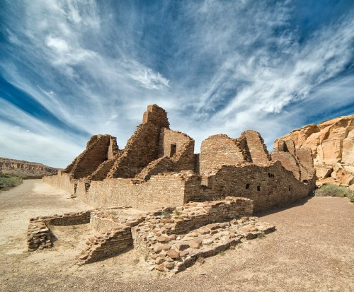 Ruins of an ancient stone structure under a blue sky with wispy clouds, surrounded by desert landscape.