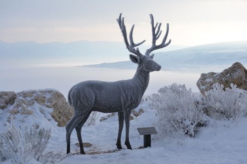 A frosted deer statue stands in a snowy landscape, surrounded by icy plants and a misty background.