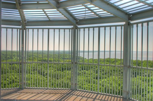 View from a lookout tower with metal railings, overlooking lush green trees and a calm body of water in the distance.