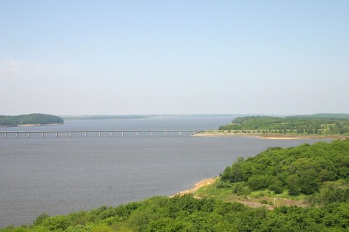 A serene view of a river with a bridge, surrounded by lush green hills under a clear blue sky.