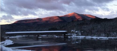 A snowy landscape with a river, a covered bridge, and mountains bathed in warm sunset light under a cloudy sky.