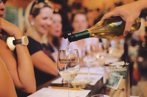 A hand pours white wine into a glass at a winery, with people enjoying a tasting in the background.