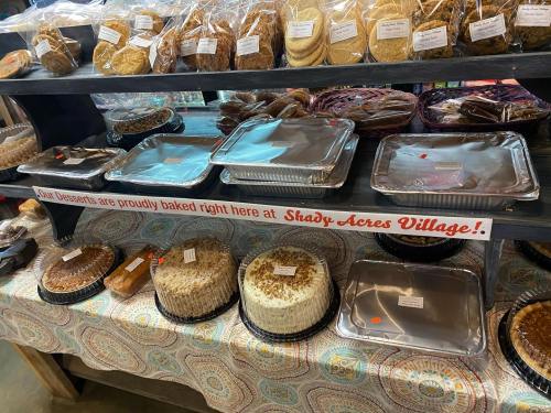 A display of various baked desserts on shelves, with a sign reading "Our Desserts are proudly baked right here at Shady Acres Village!"