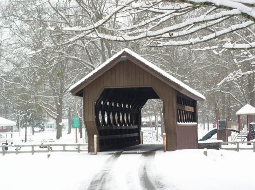 A snow-covered covered bridge surrounded by trees, with a path leading through it in a winter landscape.