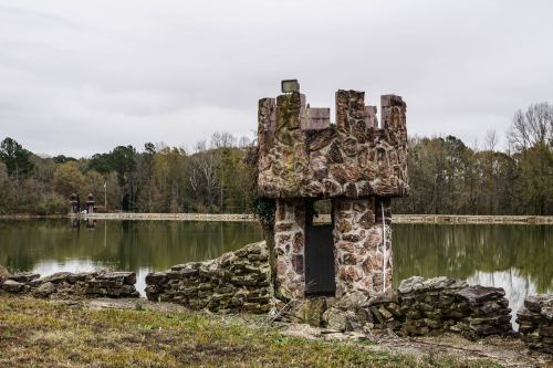 A stone tower by a calm lake, surrounded by trees and a cloudy sky, reflecting a serene, abandoned atmosphere.