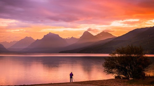 A person stands by a tranquil lake at sunset, surrounded by mountains and colorful skies.