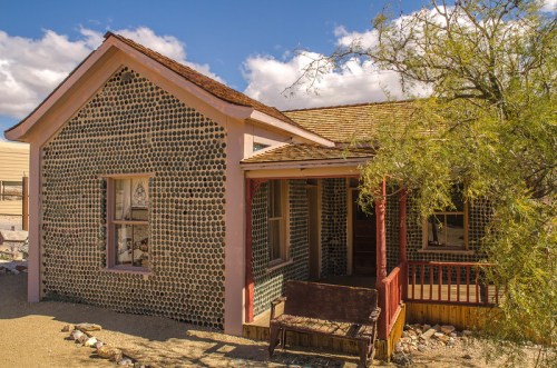 A unique house with a bottle-covered exterior, surrounded by desert landscape and a tree, under a blue sky.