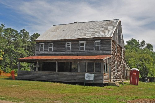 A weathered two-story wooden building with a metal roof, featuring a porch and surrounded by greenery.