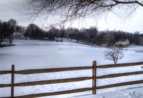 A snowy landscape with a wooden fence, trees, and distant houses under a cloudy sky.