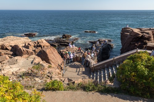 A scenic coastal view with people gathered on a rocky shore and a wooden walkway leading to the water.