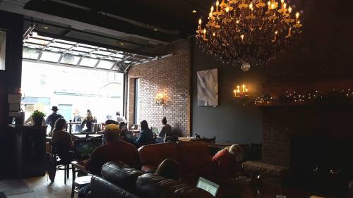 Cozy café interior with a chandelier, brick walls, and patrons enjoying coffee and conversation by an open garage door.