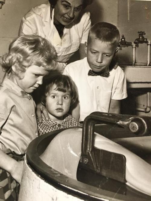 Three children and an adult woman observe a large machine, with expressions of curiosity and surprise.