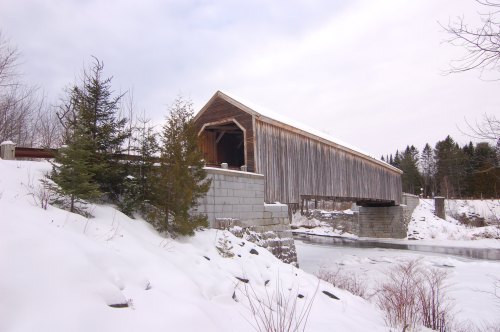A wooden covered bridge over a frozen river, surrounded by snow-covered trees and a winter landscape.