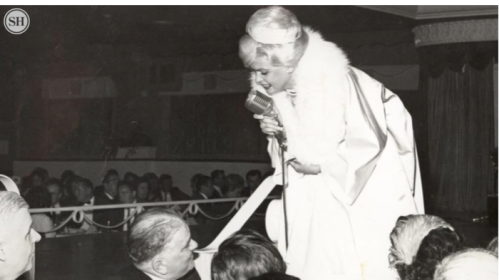 A performer in a glamorous outfit sings into a vintage microphone, engaging with an audience in a lively setting.