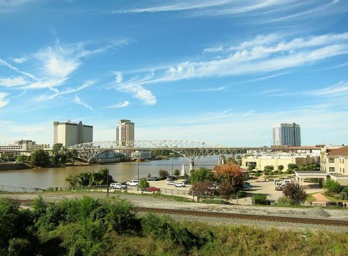 A city skyline with modern buildings, a bridge over a river, and blue skies with wispy clouds.