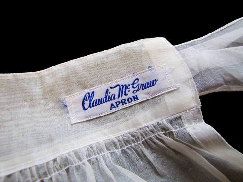 Close-up of a white apron with a blue embroidered label reading "Claudia McGraw APRON."