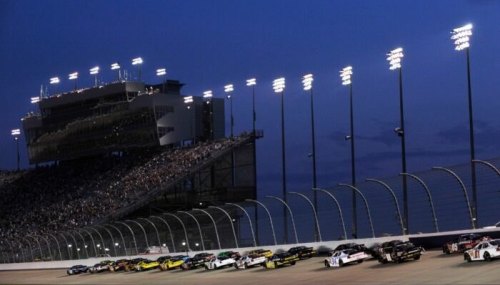 NASCAR race cars speed around a track at dusk, with a grandstand filled with spectators and bright lights overhead.