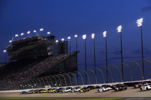 NASCAR race cars speed around a track at dusk, with a grandstand filled with spectators and bright lights overhead.