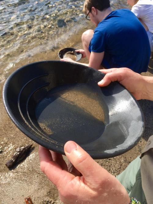 A person holds a black pan over a sandy beach, with water and sand visible inside the pan. In the background, another person is seated.