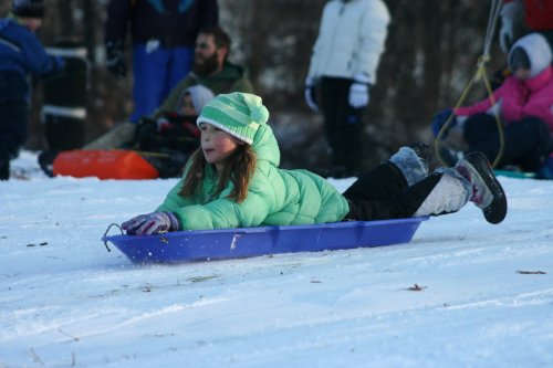 A girl in a green jacket and hat sleds down a snowy hill, enjoying a winter day with others in the background.