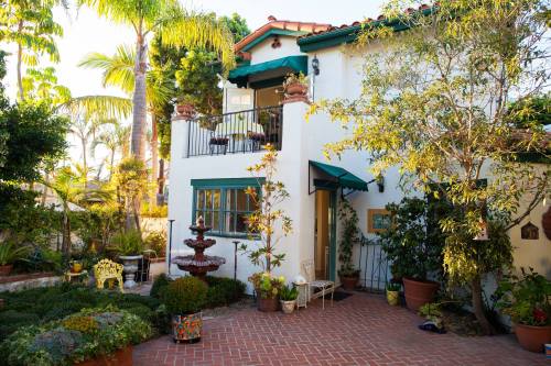 Charming white house with green accents, surrounded by lush plants and a fountain in a sunny garden setting.
