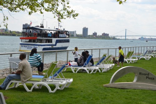 People relax on lounge chairs by the water, with a boat passing and city buildings in the background.