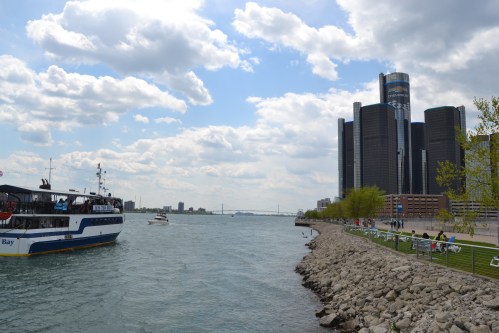 A boat on a river near a city skyline with modern buildings and a cloudy sky in the background.
