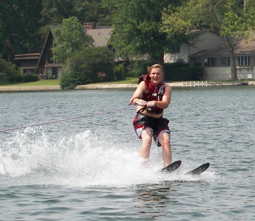 A person water skiing on a lake, creating splashes, with trees and houses in the background.