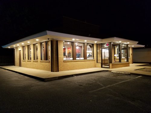 A well-lit fast-food restaurant at night, featuring large windows and a modern design, with a parking lot in front.