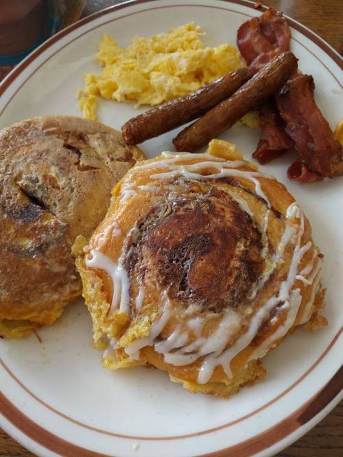 A plate with scrambled eggs, bacon, sausage links, and a cinnamon roll drizzled with icing.