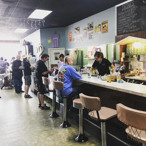 A busy diner interior with patrons at the counter and tables, featuring a menu board and retro decor.