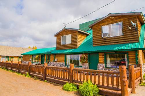 A rustic log cabin with a green metal roof, featuring a wooden fence and flower pots, set against a cloudy sky.