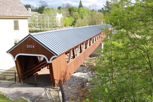 A wooden covered bridge with a metal roof spans a river, surrounded by greenery and trees. The year "2004" is visible.