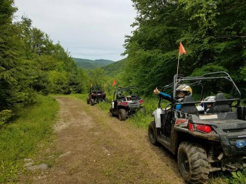 Three ATVs on a dirt trail surrounded by lush green trees, with riders giving a thumbs up.