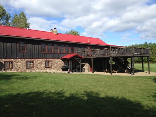 A rustic two-story building with a red roof, stone lower level, and a wooden deck overlooking a grassy area.